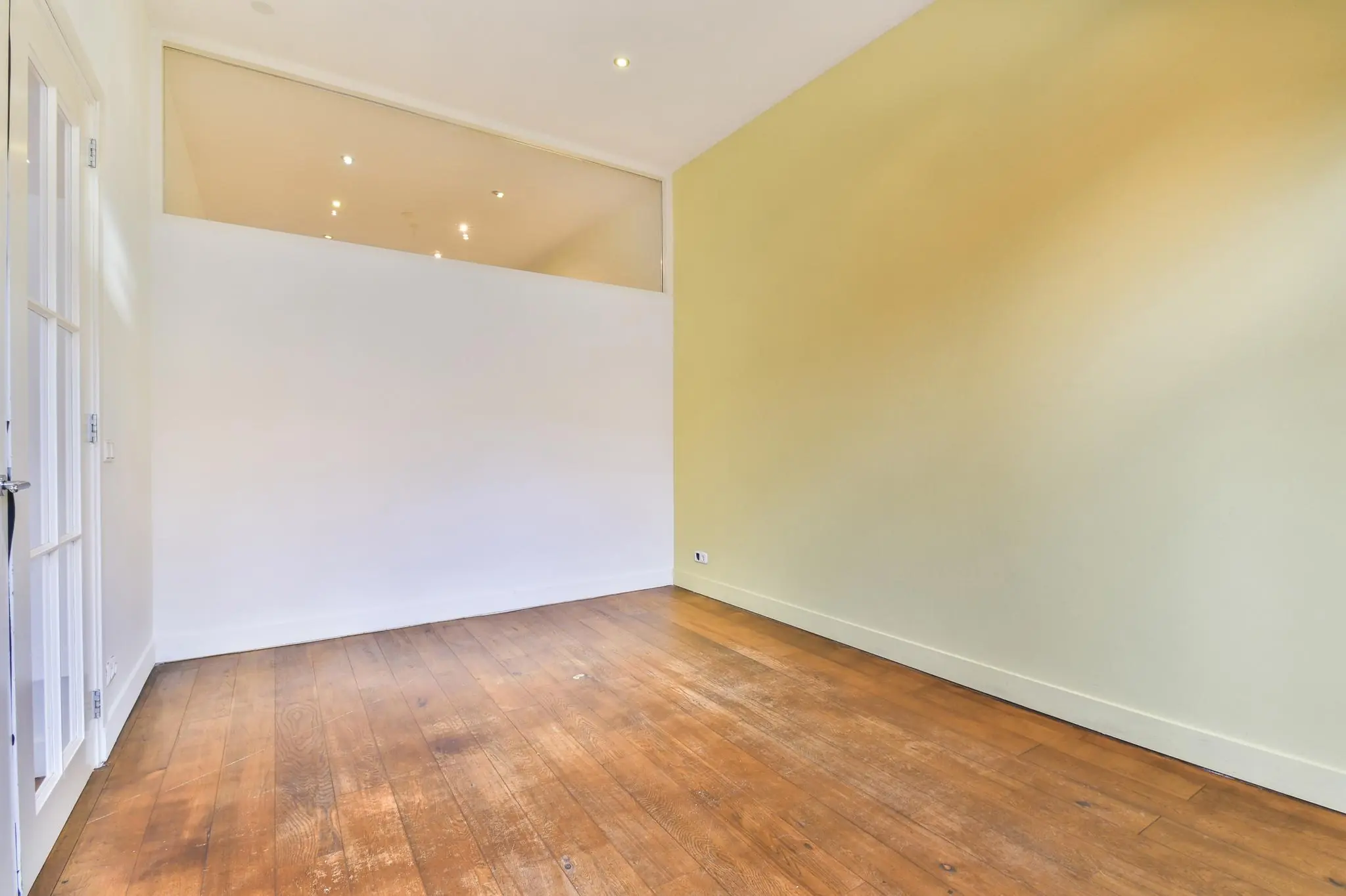 Lege kamer in Korte Schimmelstraat met houten vloer, witte en geelgroene muren en glazen bovenlicht.